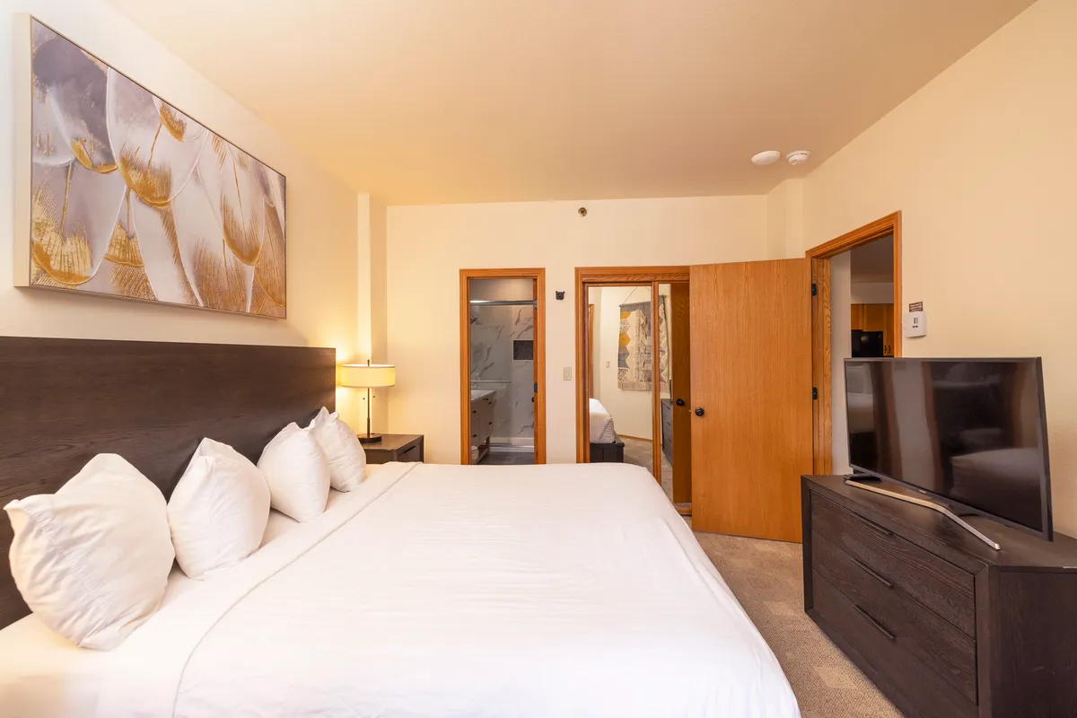 A cozy, inviting bedroom with a large bed dressed in soft white linens, decorative artwork, a wall-mounted television, and warm wooden furniture. The atmosphere is intimate and restful, ideal for couples or families seeking a peaceful night's sleep.