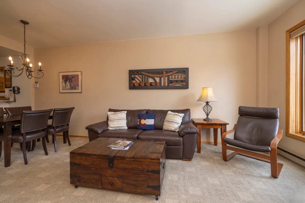 A bright, comfortable living area featuring a sofa, coffee table, dining area, and a large window that lets in natural light. The chandelier and decor create a welcoming, home-like feel.