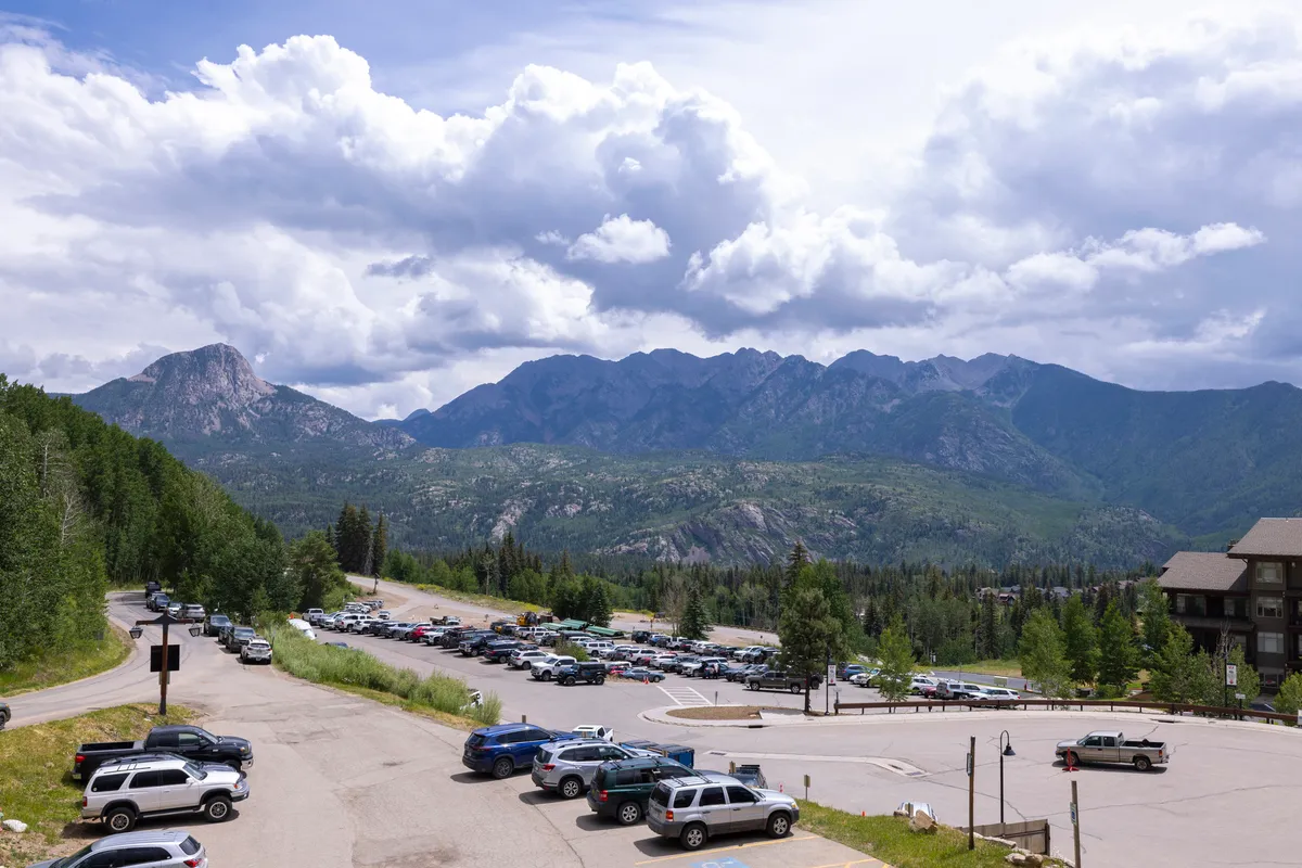 A wide-angle view of the majestic mountain range under a dramatic, cloudy sky. The foreground includes a parking lot and is framed by lush surrounding trees, hinting at immediate access to nature and panoramic vistas.