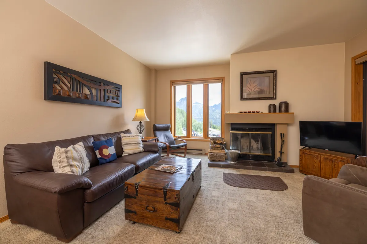 A warm, inviting living room features a plush sofa with colorful cushions, a wooden coffee table, and an armchair arranged around a cozy fireplace. Large windows fill the room with natural light and showcase stunning mountain views, seamlessly connecting the indoors to the outdoors.