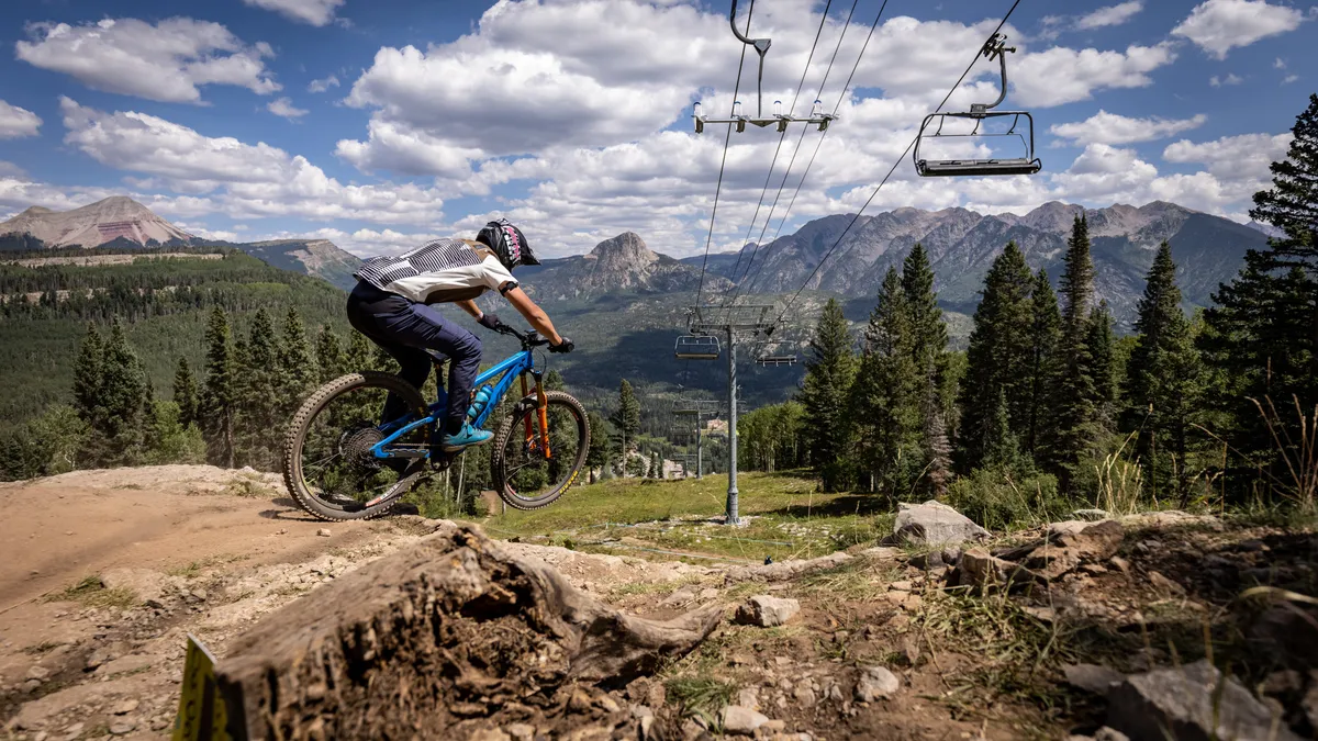 An energetic outdoor scene showing a mountain biker on a dirt trail, a ski lift in operation, panoramic mountain views, lush trees, and an inviting sense of adventure. The image reflects year-round recreational opportunities, from mountain biking and hiking to scenic chairlift rides.