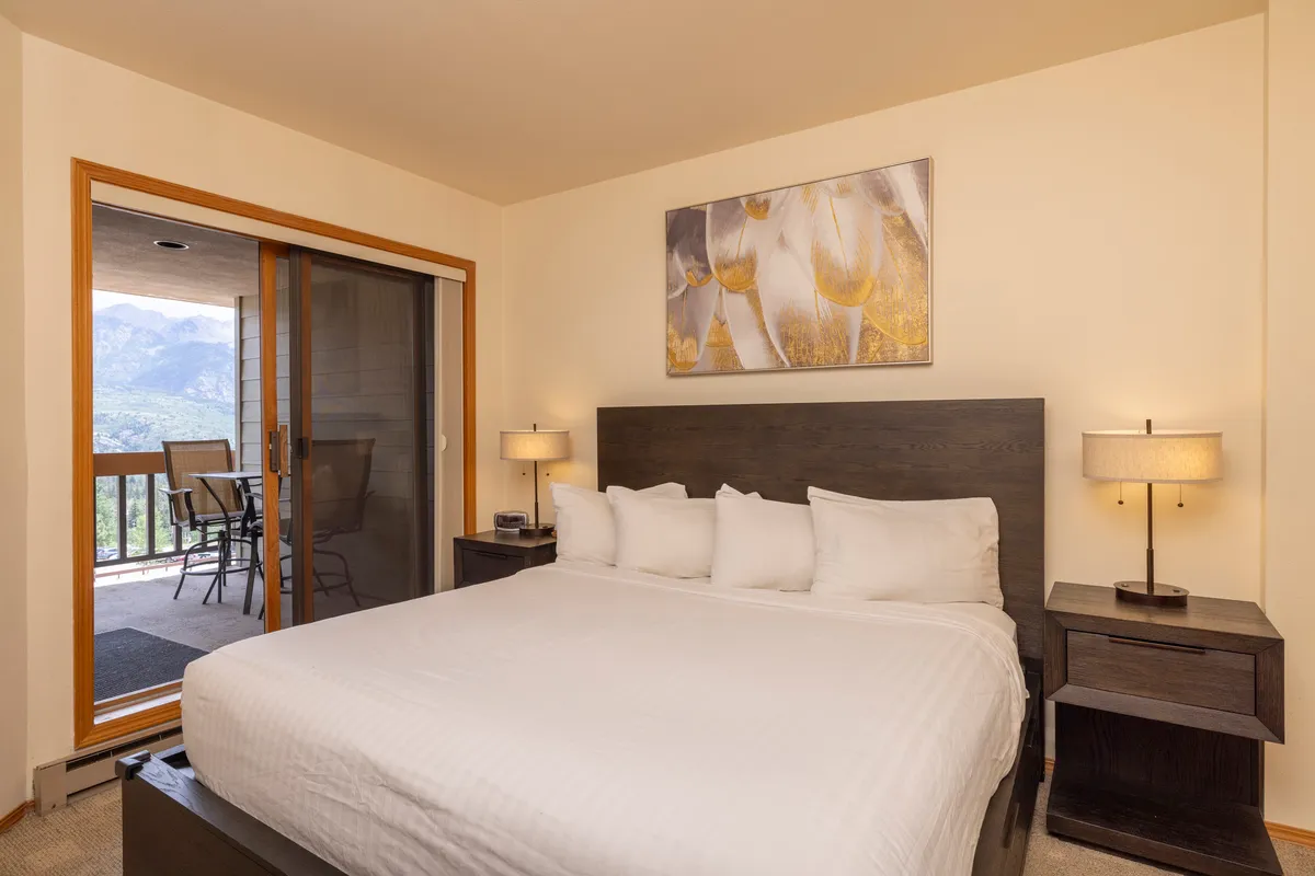 A serene bedroom with a crisp white king-size bed, tasteful framed artwork above the headboard, and a sliding glass door that opens onto a private balcony. The space is bathed in soft natural light, accentuating the tranquil ambiance, perfect for relaxation and privacy.