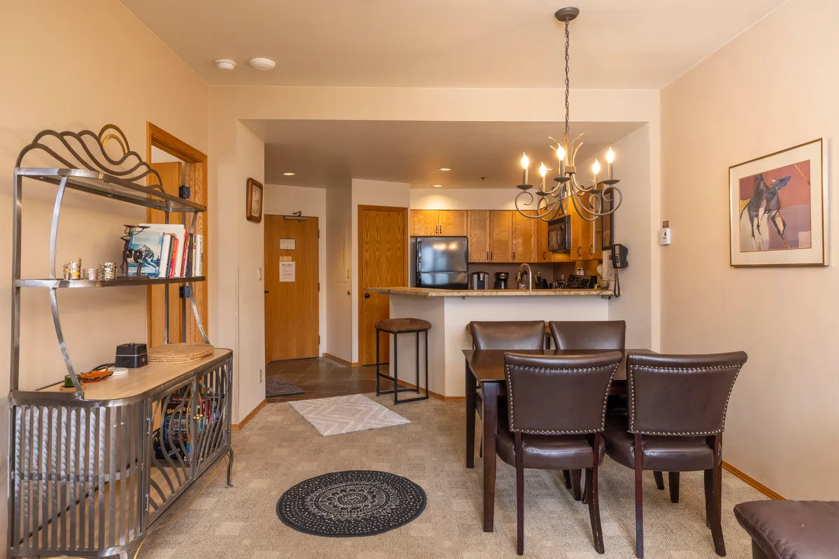 The image features a dining table under a chandelier, with a view into the open kitchen area and a decorative shelf in the background. The space is styled to encourage relaxed family meals and entertaining guests.