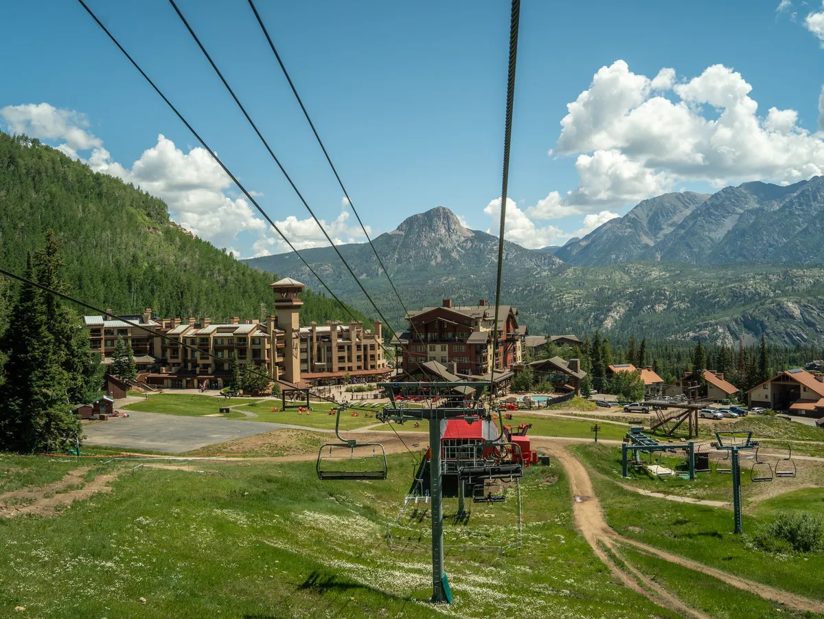 A panoramic shot featuring a ski lift ascending toward a stunning mountain range, with lodge buildings nestled among the green landscape. The composition evokes outdoor adventure and inviting village charm.