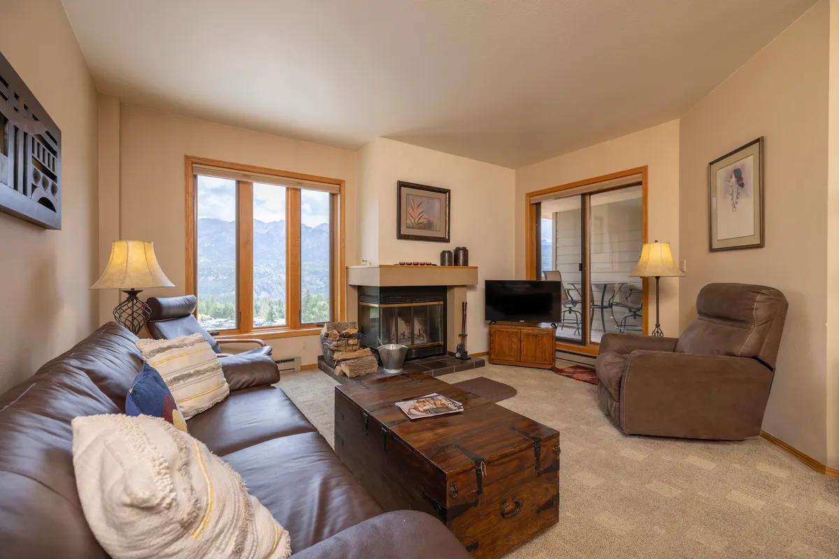 A serene living space featuring a comfortable leather sofa, a wooden coffee table, a glowing fireplace, and large windows that offer panoramic mountain views. Natural light floods the room, creating a welcoming environment for relaxation or socializing.