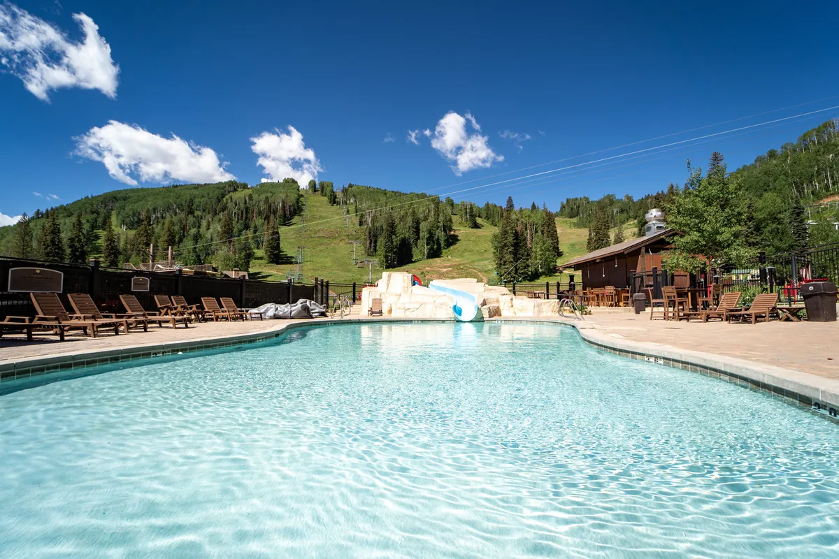 A cheerful scene featuring a sparkling swimming pool with a water slide, lounge chairs, wooden deck, and a dramatic mountain backdrop. Families are shown relaxing and socializing poolside, with scenic views reinforcing the resort’s recreational amenities.