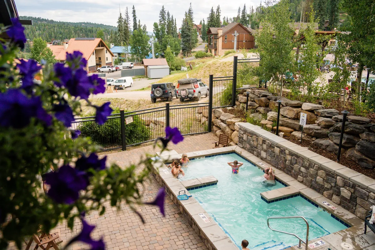 An inviting outdoor hot tub surrounded by blooming purple flowers and towering pine trees, with nearby cozy cabins. The scene radiates warmth, relaxation, and a social yet intimate setting perfect for couples.