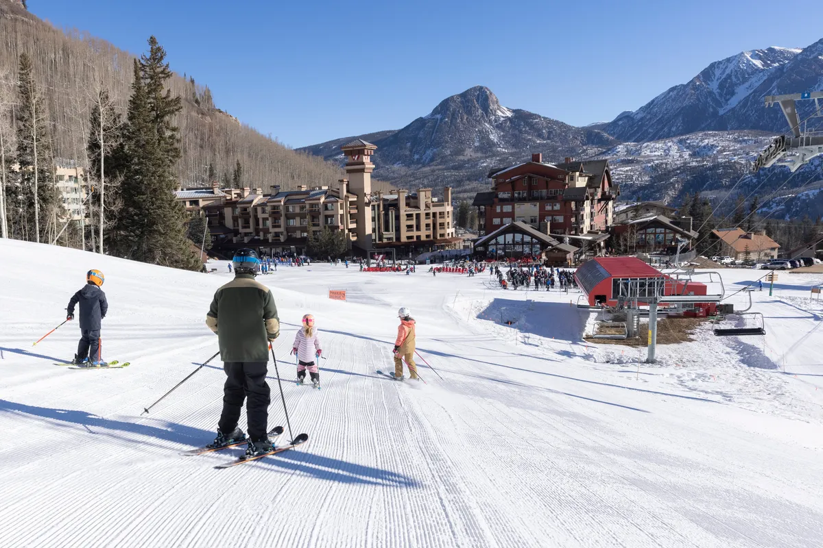 This image captures skiers gliding down pristine slopes with a dramatic mountain range serving as the backdrop. The foreground highlights active visitors enjoying the snow, while ski lifts and resort buildings create a lively, adventure-filled atmosphere.