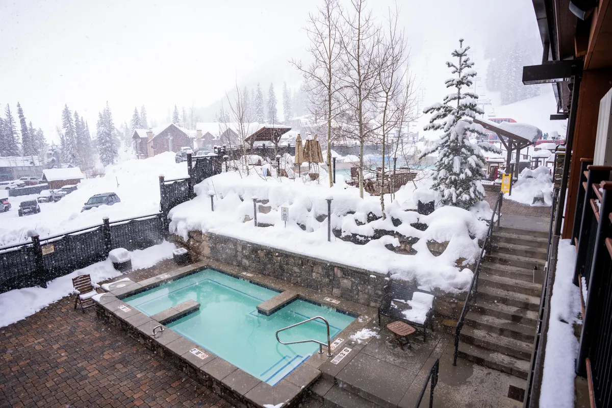 A snow-covered landscape frames an inviting outdoor hot tub nestled among evergreen trees and wooden structures. The scene is tranquil, with steam rising from the hot water and hints of social gathering or relaxation after a day on the slopes.