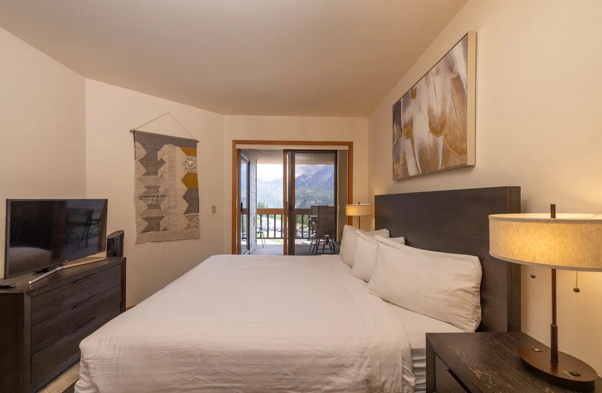 A cozy bedroom with a plush bed, tasteful artwork, and a balcony offering expansive views of the surrounding mountain landscape. The room exudes calm and privacy, with the outdoors just a step away.