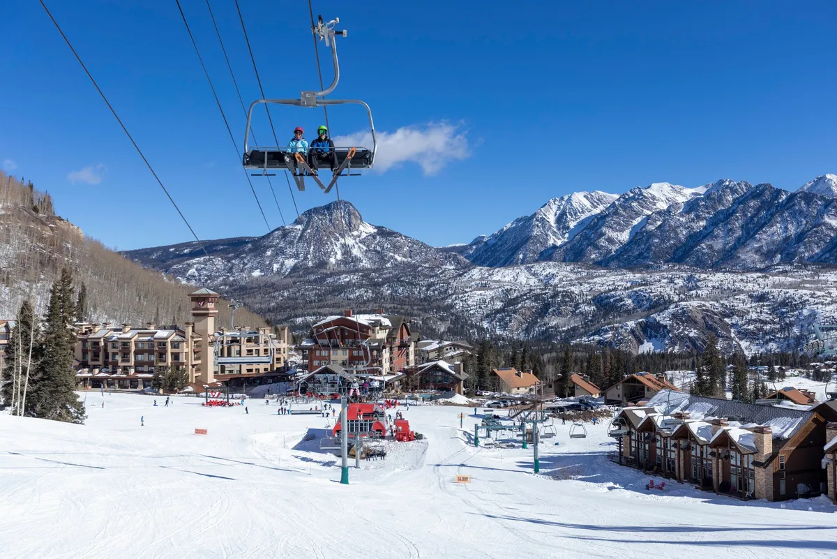An exterior scene of Purgatory Village with ski lifts, snow-covered mountains, skiers on the slopes, and lodging buildings. The area is lively with people enjoying outdoor activities and the mountain ambiance.